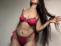 Tisia with long brown hair, wearing pink lace lingerie, her tits covered, showing her pussy, holding hair, pale skin, standing, slightly leaning forward, soft lighting.