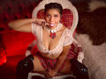 LucieMilner with brown skin, braided hair, lace top, plaid skirt, black boots, and pink ball gag, sitting on a white chair, showing her boobs, and holding her gag.