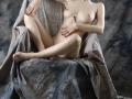 Nude KamiLove with small tits, pale skin, and red nails, sitting on a chair with gray drapery, legs crossed, hand covering her pussy, other hand on her ass.
