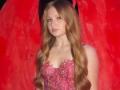AkilahGhil has long, wavy blonde hair, wearing a pink, sequined, strapless dress, with small, round tits, and a silver necklace, against a red backdrop.