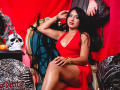LilyConorr in a red dress, showing big tits and a cute ass, sits on an ornate chair, black hair, gold bracelet, hand on head, skull and pumpkin on left.