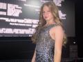 AkilahGhil with long blonde hair, wearing a sheer black dress with silver sequins, standing, smiling, showing her tits through the fabric, in a dimly lit room.