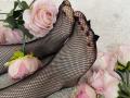 MiraRosse's feet in black fishnet stockings, with pink and white roses scattered around, showing slightly dirty soles with visible black marks. Soft white furry background.
