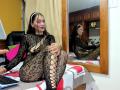 AfroditaHan with long black hair, wearing a black lace bodysuit with large cutouts, sitting on a bed, reflecting in a mirror, laptop on the bed, smiling.