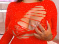 Genexis wearing a red, cut-out, sparkly dress with large slits, exposing her red lace bra-covered tits. Her hand with tattoos is on her chest, showing painted nails.