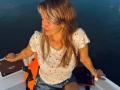 MikyWayxy with long blonde hair, white shirt with small prints, denim shorts, orange life vest, sitting on boat, smiling, right hand on boat edge, left arm resting.