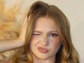 AkilahGhil has long, wavy blonde hair, one eye closed in a wink, and is making a pouty, pursed-lip expression. She's touching her head with her right hand.