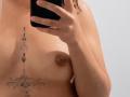 EmilyEvan's topless selfie shows her holding a phone, with her small brown tits and a tattoo between her breasts. Her right arm is raised, covering part of her face.