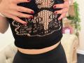 Stefanni's hands with red nails cover her black mesh lace top, revealing her bare boobs through the pattern. She's wearing black pants, in a cozy indoor setting.