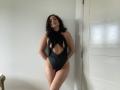 Dionisia with dark wavy hair, wearing a black cutout one-piece, showing her tits and pussy, standing against a white door, hands behind her back, looking sexy.
