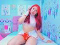 MiaLeroy with red hair, wearing a white crop top, red and white miniskirt, and red panties, sitting on a bed, hand touching her hair, showing her tits and pussy.