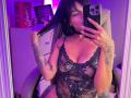 MajoJones with black lace lingerie, holding phone in mirror selfie, showing tattoos, holding hair, purple lighting, large boobs, lace-up front, chair, holding phone with black case.