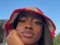 JenifferStar with dark brown skin, wearing a red and beige straw bucket hat, nose ring, and pink lipstick, resting her chin on her hand, under a blue sky.