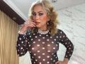 MisseScarleth with blonde curls, wearing a sheer black polka dot top, showing her tits through the fabric. She's got a pearl necklace, red lipstick, and a ring on her finger.