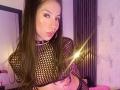 NahomyCarther with long brown hair, wearing a black fishnet top, showing her tits, with a nose ring and a glowing light, pouting her lips, in a purple-lit room.