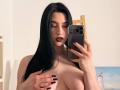 MaryLori with long black hair, dark lipstick, and black nail polish, holding a phone, covering her large tits with one hand, taking a mirror selfie.