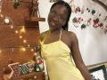 LilNaughtySally with dark skin, braided hair, glasses, and yellow dress, standing next to a wooden Christmas tree with lights, ornaments, and a Santa doll.