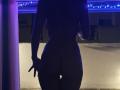 Silhouette of JuneLaFrenchie, standing naked, with visible ass and hand making a peace sign, under blue LED strip lights, in a dimly lit room.