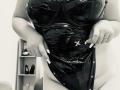 DiLaria, vêtue de lingerie en latex noir brillante, tire le devant vers le bas pour exposer sa peau douce et pâle, ainsi que ses seins volumineux et ronds. Ses longs ongles soignés et sa bague sont visibles..