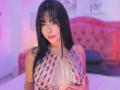 GraceClarke with long black hair, wearing a sparkly chain top, exposing her large tits, standing in a pink-lit bedroom, looking at the camera with a slight smile.