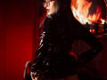 TatsumiLee with dark blue hair, glasses, and black latex outfit, standing, looking back, hand on hip, holding a gun, fiery red background, butt facing camera, sexy.