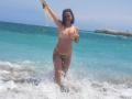 LauraSin is standing in the ocean, topless with small tits, wearing a green and yellow bikini bottom, holding her top in the air, and smiling. Water splashes around her.