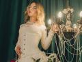 KylaHemeke with blonde wavy hair, wearing a white corset dress, holding a fancy candelabra with pearls, glowing lights, and green velvet curtain background. Boobs visible under dress.