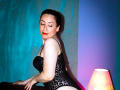 SweettEve with pale skin, dark hair, red lipstick, black lace corset, leaning on a table, smiling, next to a lamp with pink and blue lighting. Boobs, ass, and tits visible.