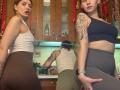 Three HotCaramelsTrio women in a kitchen, wearing sports bras and tight pants, showcasing their asses and tattoos. One has a white bra, another a blue bra, and the third is facing away.