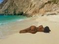 Berrie69 lies face down on a sandy beach, her back to the viewer. She has dark brown skin and a curvy, well-toned body. She wears a minimalistic string bikini, highlighting her round buttocks. Her long black hair is slightly messy. The beach has fine, white sand and clear turquoise water. A rocky, beige cliff rises in the background, contrasting with the bright blue sky. The sunlight casts subtle shadows, emphasizing her body's contours. The image focuses on the natural beauty of the beach and Berrie6.