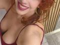 Khharha, con i capelli rossi, sorridente, che indossa un rossetto rosa scuro e una maglia color borgogna a scollo basso, che mostra il suo seno prosperoso, orecchini a cerchio dorati e un piccolo tatuaggio sulla spalla..