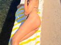 MiraGi lying on a yellow and white striped towel, wearing a black and white polka dot bikini, with her ass and back to the camera, on a sandy beach.