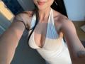 Olieri in a white halter dress with deep cleavage, showing her tits and a small tattoo on her chest. She has long black hair and a slight smile.