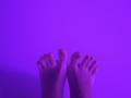 MargoFun's feet are up in the air, toes slightly curled, against a solid purple background. Her toenails are painted a light orange.