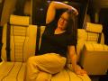 BadAngel with glasses, black top, beige pants, sitting in a cream leather SUV, arm raised, hand in hair, smiling, medium tits, brown hair, red nails, relaxed.