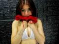 LilithStorm with red bra pulled up to her mouth, biting it, showing her tits in a white bra, tattoo on left arm, dark brown hair, black brick wall.
