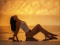 VirgieCrotts is sitting on a reflective surface, back arched, legs stretched out, wearing a white one-piece, her ass and tits visible, against a golden sunset.