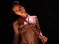 TriciaBarnes with her tits bound in rope, holding the rope with her hands, has a tattoo under her tits and a belly button piercing, in a dark setting.