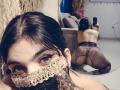 AniisaAli, wearing lace mask, fishnet stockings, and no top, on knees, facing mirror, showing her ass. Reflection shows her taking a selfie with her phone.