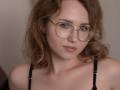 TeresaBirt with wavy brown hair, wearing glasses, and a black bra, has blue eyes, a cute face, and a neutral expression. Her tits are small and her pussy is unseen.