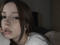 TaniaRaffa with brown hair, white shirt, and pouty lips, lying on bed, showing her white lace panties and butt. She has dark eyeliner and a finger near her mouth.