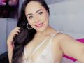 CamilaBardot with long black hair, smiling, wearing white lace lingerie, showing her tits, earrings, red bracelet, and pink lipstick, in a bedroom selfie.