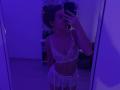 SerenaHuseth in purple-lit mirror selfie wearing white lace lingerie, showing her small tits and flat ass. She's holding a phone, her hair in a messy bun.
