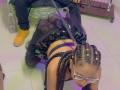 LilithAndAsmodeo, on knees, wearing bra, black dress, and glasses, with braided hair, being held down by a guy in yellow sneakers, his cocky hand on her back.