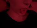 DarylSanks with red-lit skin, sticking out tongue with a pierced lip, black shirt, and visible ear studs, partially shadowed, showing a slightly mischievous expression.