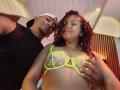 LilithAndAsmodeo with curly hair, wearing neon yellow lace bra, exposing her tits, being held by a black-capped man in a black shirt, looking down, in a dim room.