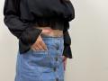 Tilliana with a black sheer top, blue button-up skirt, and pink nail polish, showing her bare tan belly and hand in a pocket.