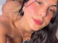 SarahPrincess with pink blush, red lipstick, and long black hair, looking at the camera, topless, showing her bare shoulders and chest, with a silver necklace, lying down.
