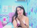 MiaLeroy with long black hair, white headband, fishnet arm, pink nipple pasties, smiling, hand on chin, light blue room, dreamcatchers, air conditioner, pink headboard, light skin.