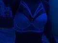 A close-up of ShylaCatillo's chest, illuminated in blue light. She's wearing a white, strappy bra that accentuates her large breasts. The shadowy room adds a sensual, mysterious vibe.